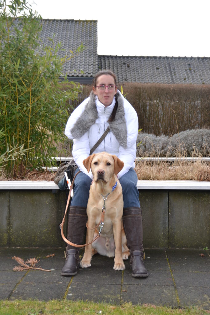Foto van Debby die, tijdens de schoolinstructie van februari 2012, op een muurtje zit bij KNGF Geleidehonden met blonde labrador Tobias bij haar voeten.