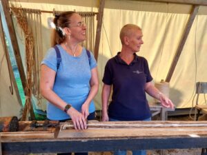 Foto van een vrouwelijke touwslager (rechts) en Debby (links) die onder een luifel staan. Debby kijkt naar de touwslager en houdt haar handen op een houten werkbank. Op de achtergrond zijn verschillende stukken touw te zien, die aan houten staven zijn bevestigd. Er staan ook nog enkele gereedschappen en materialen op de werkbank.