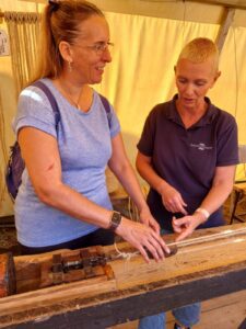 Foto van een vrouwelijke touwslager (rechts) en Debby (links), die samen aan een houten werkbank / lijnbaan staan om touw te maken. OP de werkbak is een mechanisme zichtbaar. Ze staan buiten onder een luifel. Op de achtergrond zijn enkele andere gereedschappen en mogelijk bundels koord of touw te zien.